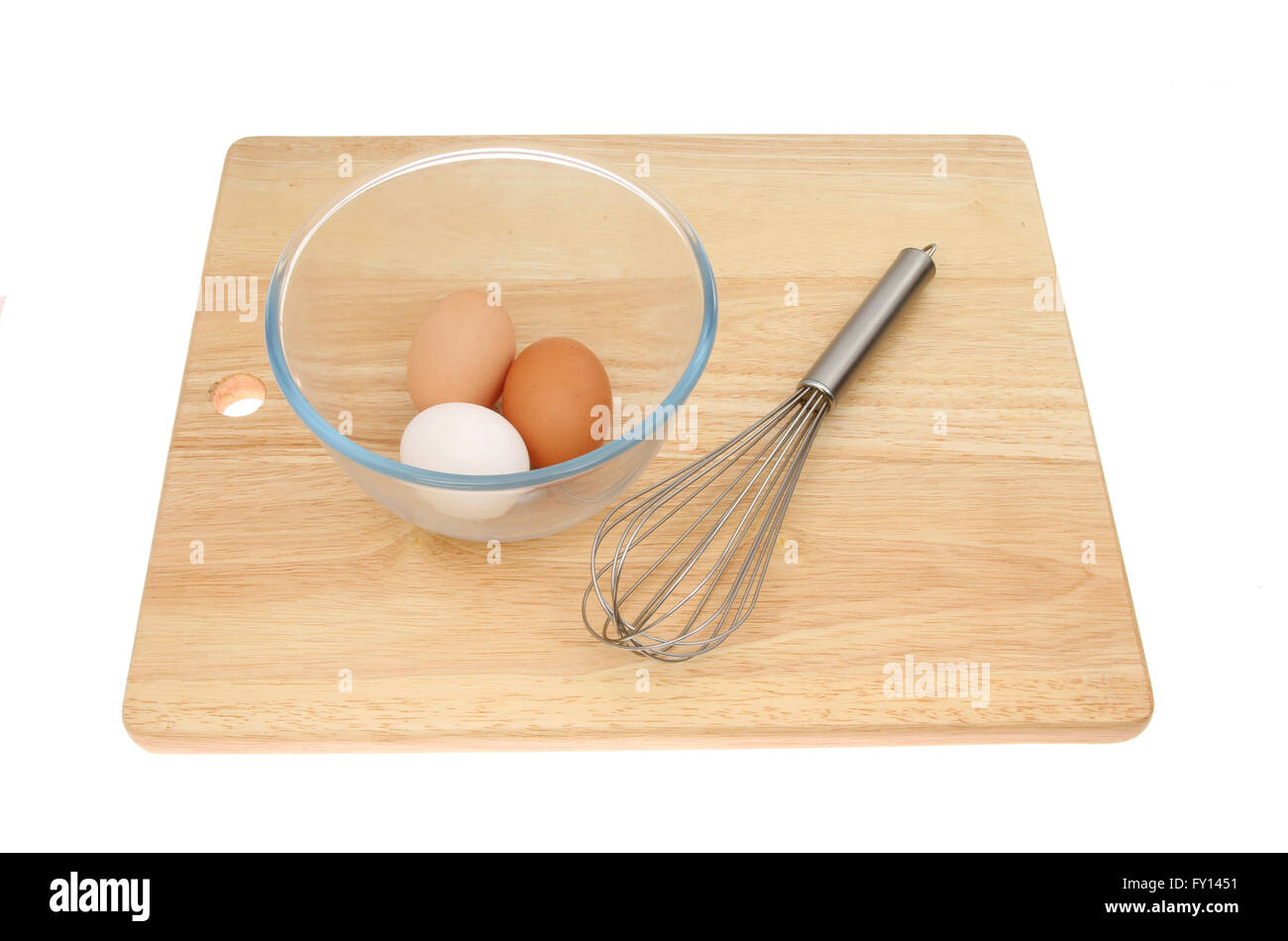 Three eggs in a glass mixing bowl with a whisk on a wooden chopping board isolated against white Stock Photo