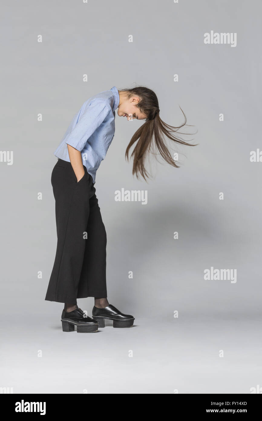 Smiling woman with tousled hair and hands in pockets standing against gray background Stock Photo