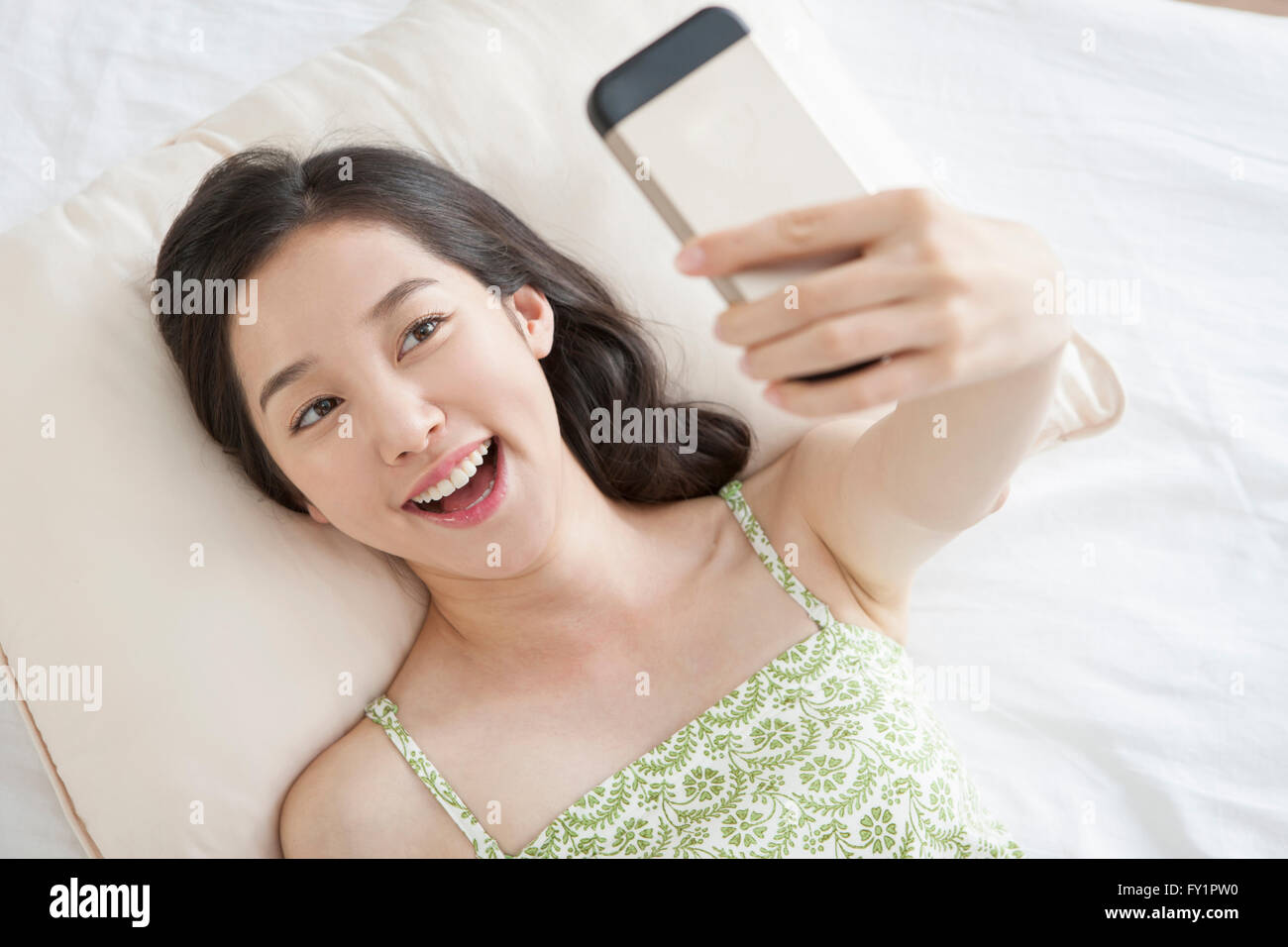 Portrait of young smiling woman lying down on bed and taking a picture of herself with smartphone Stock Photo