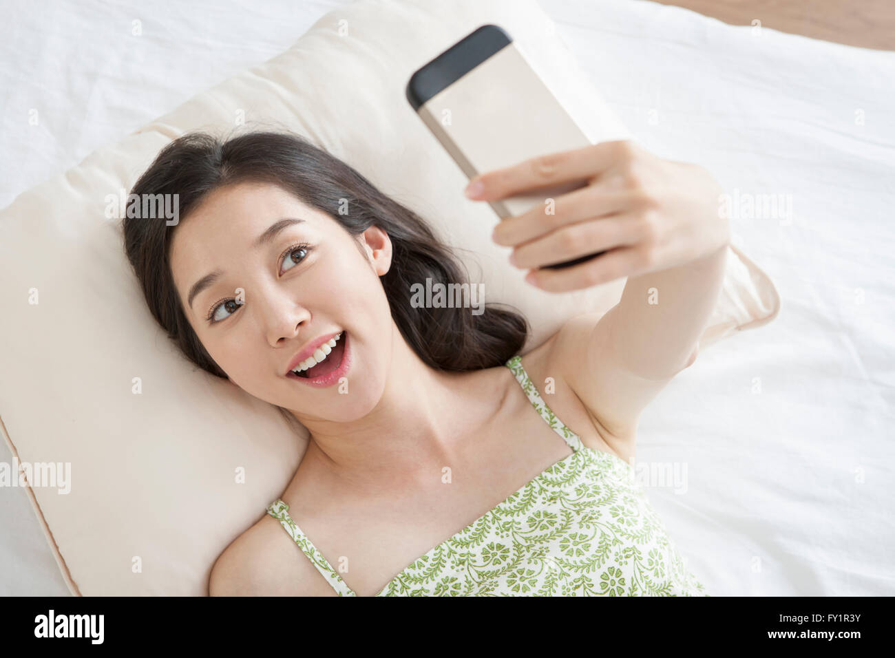Portrait of young smiling woman lying down on bed and taking picture of herself with smartphone Stock Photo
