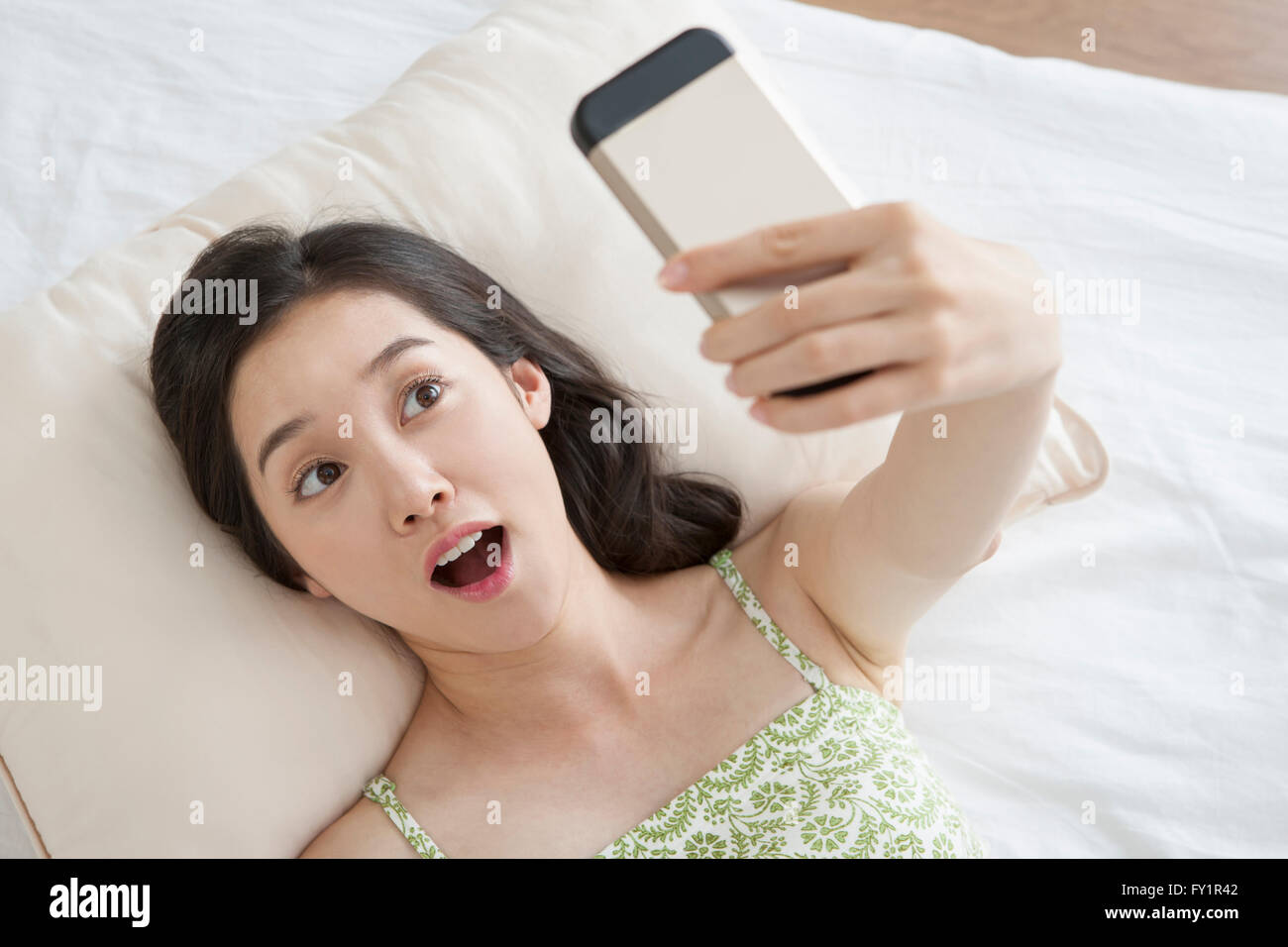 Portrait of young smiling woman lying down on bed taking picture of herself looking up Stock Photo