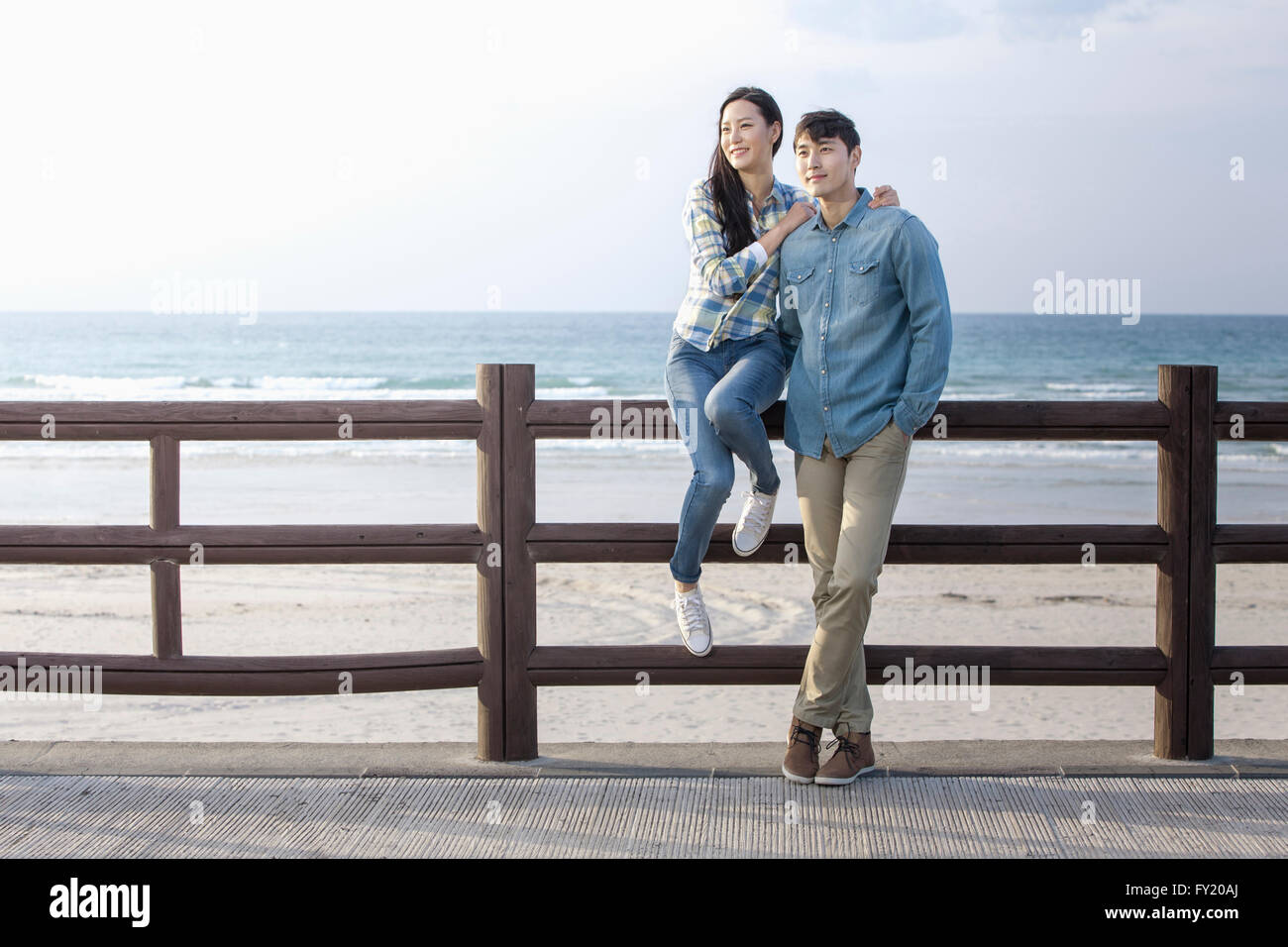Woman sitting on a fence and a man leaning his body on a fence along the beach Stock Photo