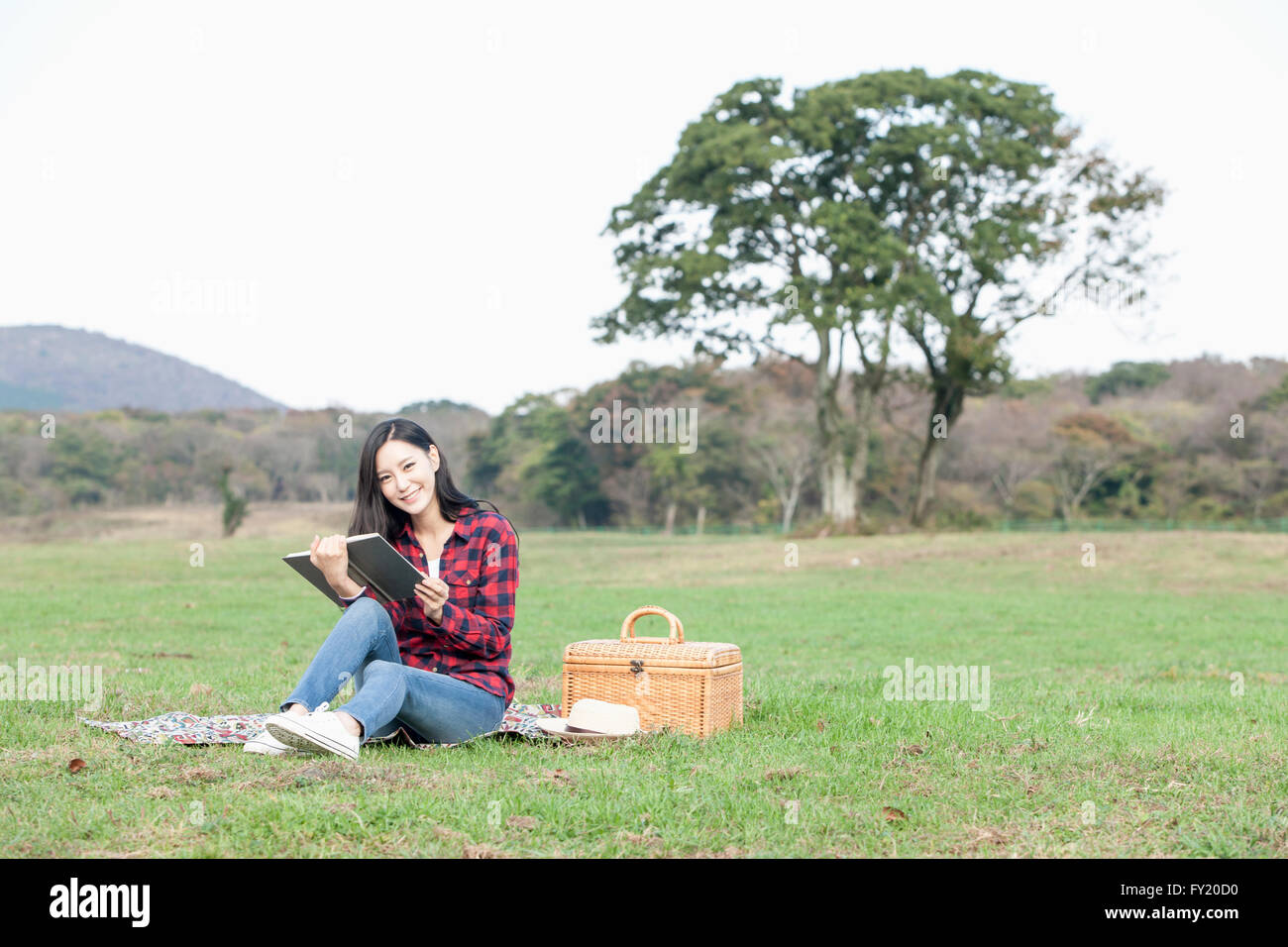 Woman seated on grass and holding a book with a picnic box at the field Stock Photo