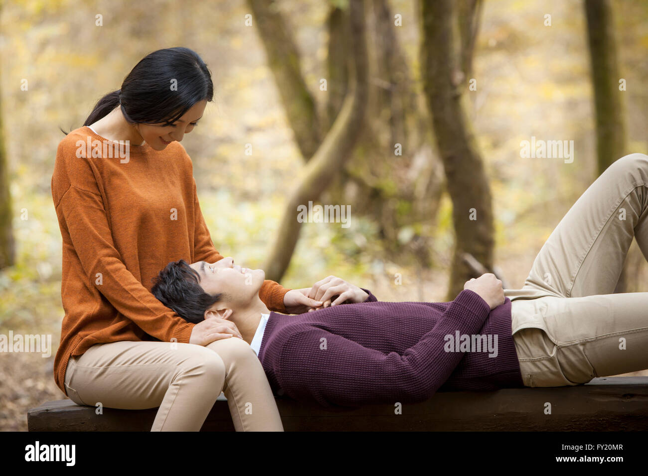 Woman seated on a bench and a man lying on her lap looking at her at the forest in fall Stock Photo