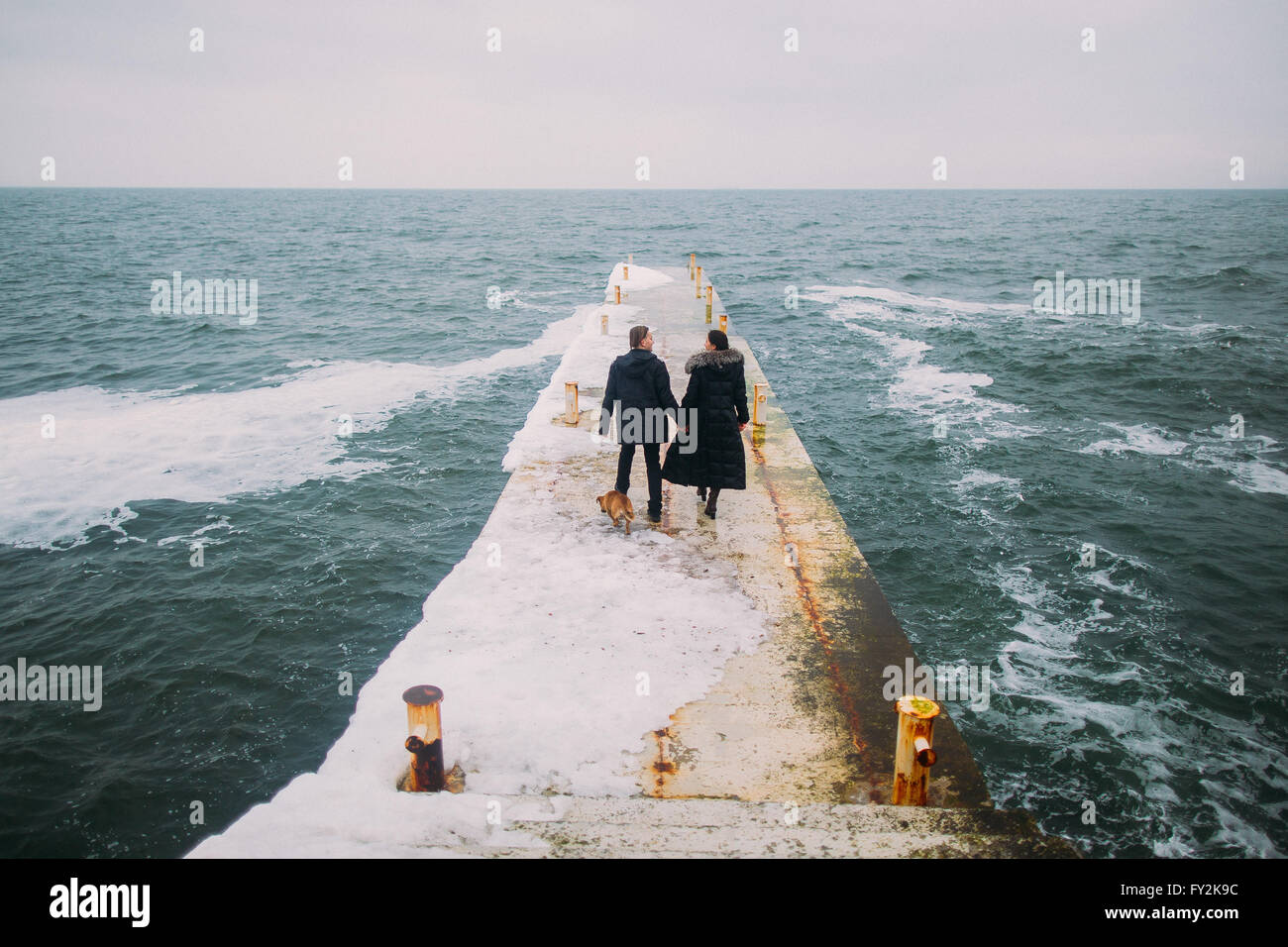 Rear romantic view of a young couple walking together on stoned pier during rainy autumn day. Winter sea background Stock Photo