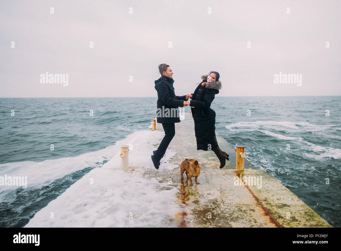 Rear romantic view of a young couple walking together with little dog and having fun on stoned pier during rainy autumn day. Winter sea background Stock Photo
