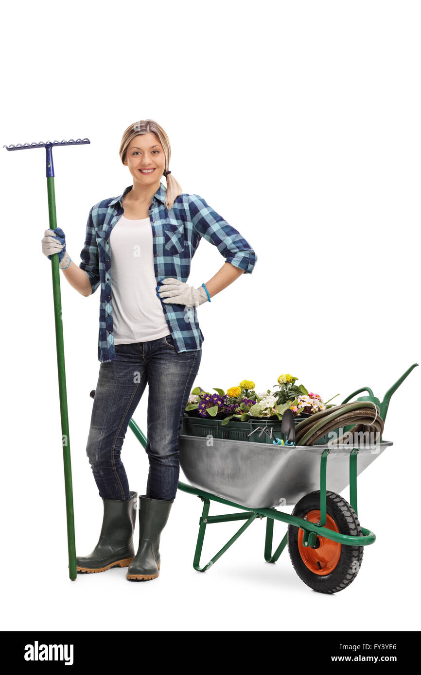 Full length portrait of a woman holding a rake and posing next to a wheelbarrow full of gardening equipment and flowers isolated Stock Photo
