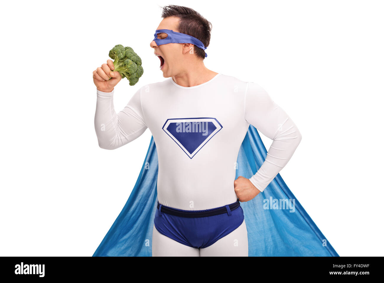 Young guy in a superhero outfit taking a bite of a large piece of broccoli isolated on white background Stock Photo