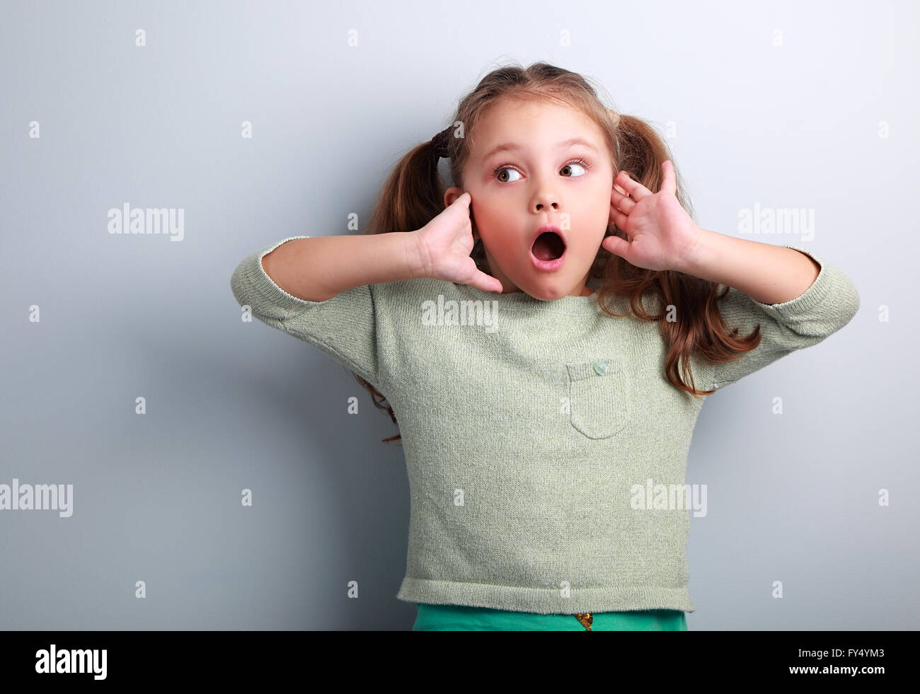 Surprising kid girl with long hair with opened mouth and hand near face looking on blue copy space background Stock Photo