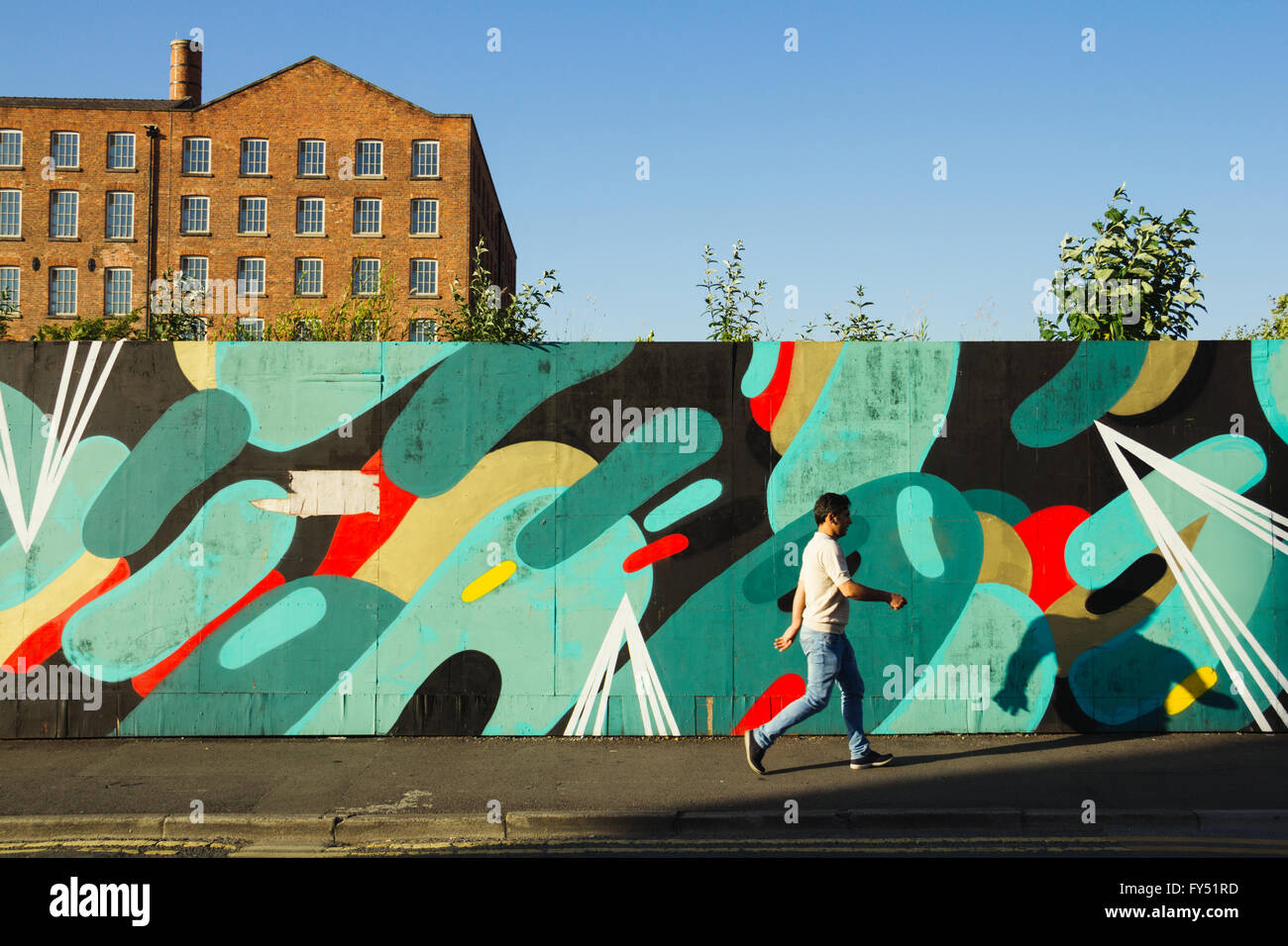 Man walking by a piece of street art in Manchester, with long shadow and mill in the background. Stock Photo