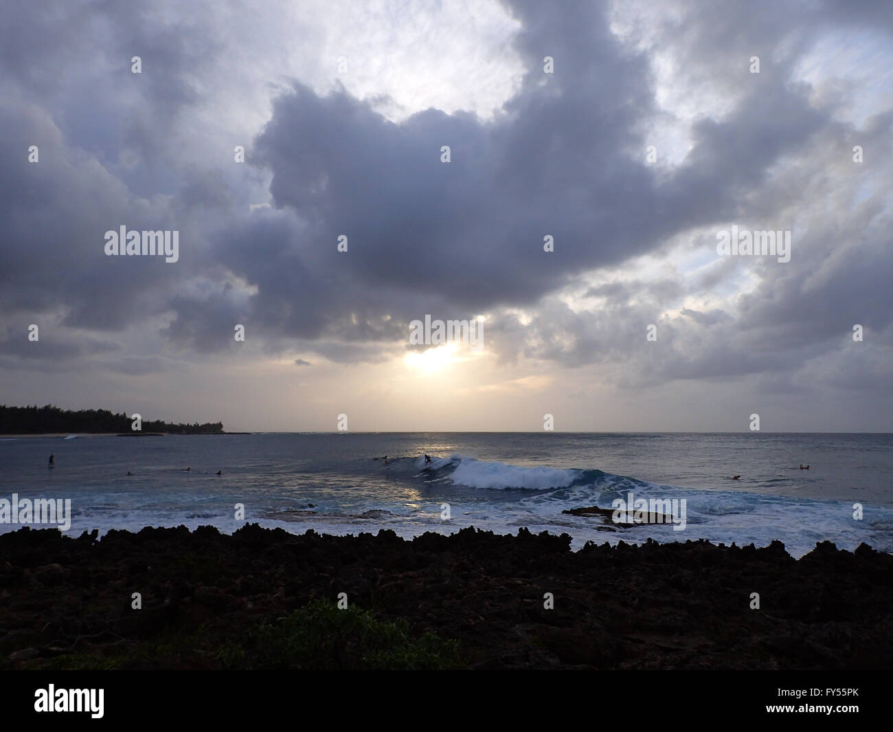 Surfers ride waves as sunsets though the clouds off the coast of Turtle Bay on the North Shore of Oahu, Hawaii. Stock Photo