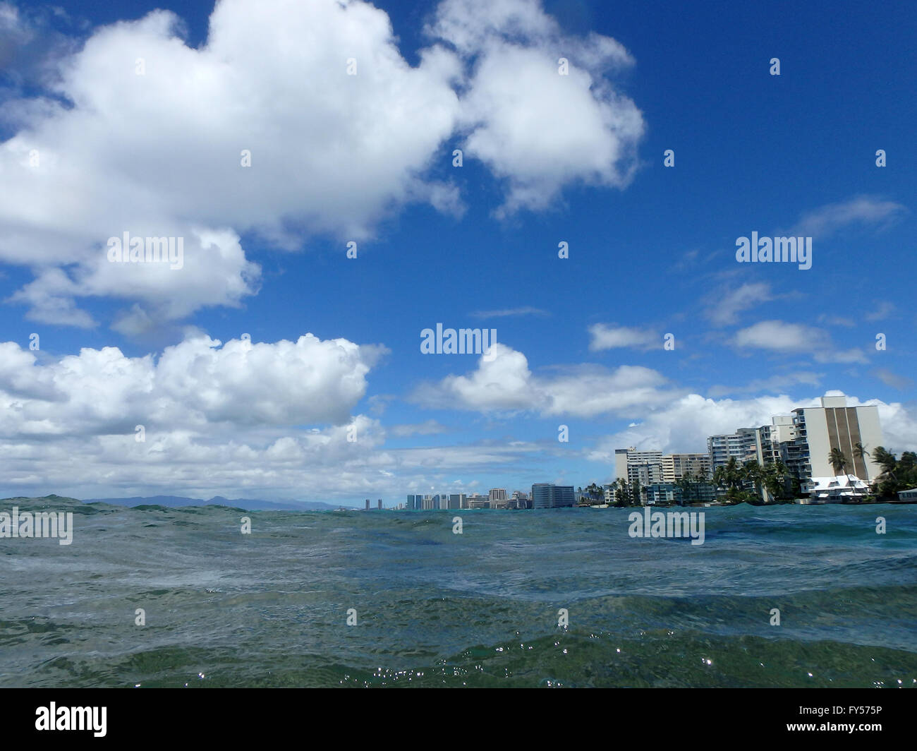 Ocean waters ripple off the coast of waikiki with hotels lining the water and clouds in the sky on Oahu. Stock Photo