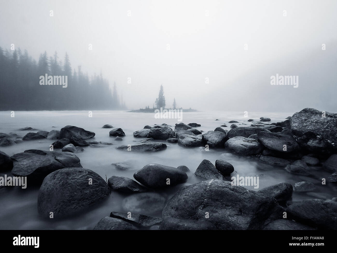 A moody monochrome image with rocks in the foreground and an island with a solitary tree in the background against a misty sky. Stock Photo