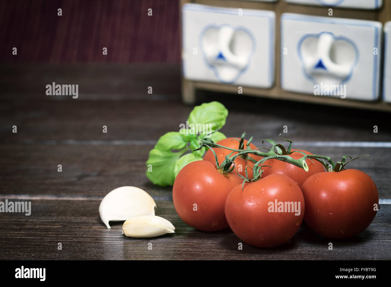 Vintage photo of cherry tomatoes, basil and garlic lying on the wooden table. Porcelain spice rack is in the background. Stock Photo