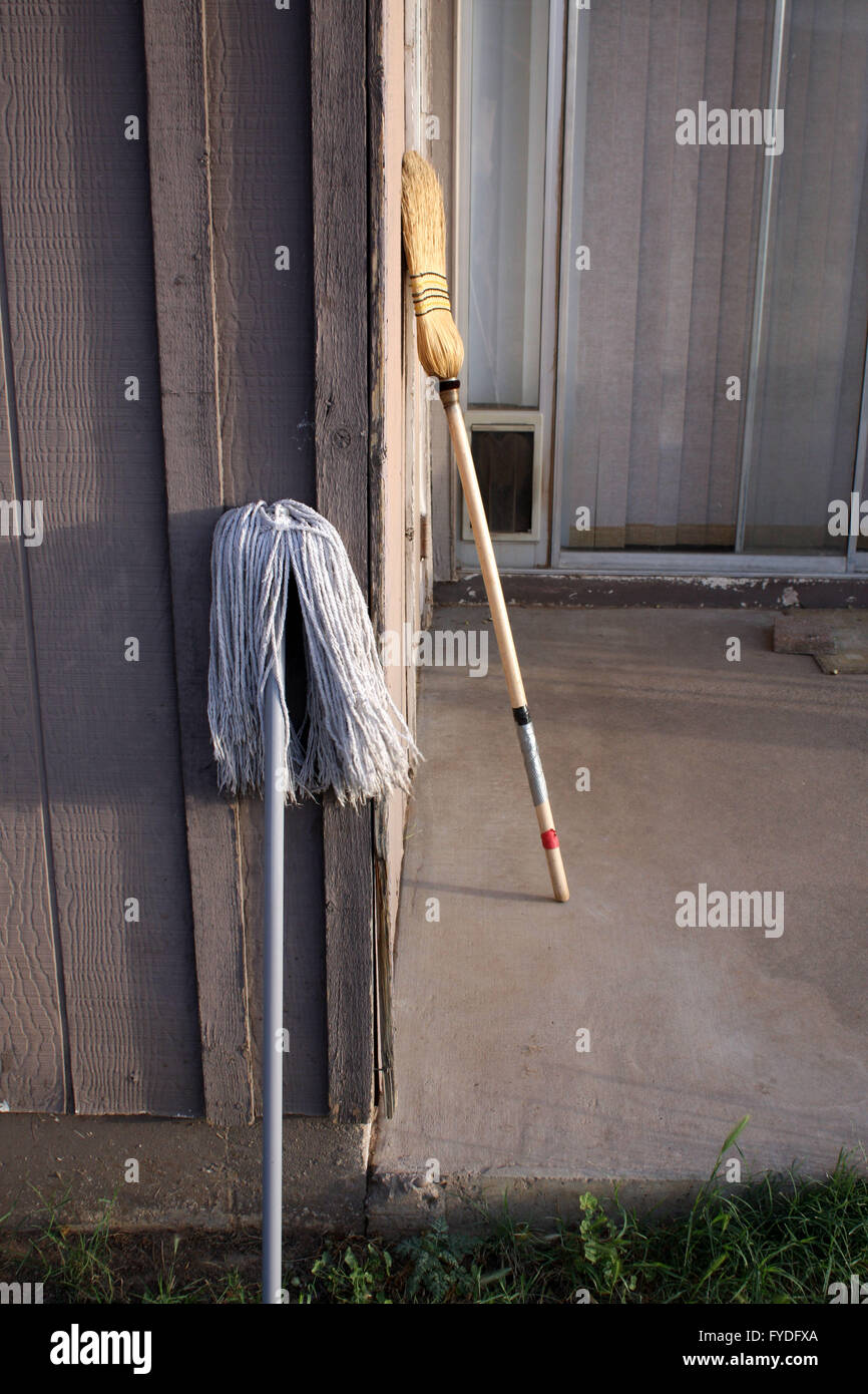 A yellow straw whisk broom and a Grey mop lazing up against the back of the house at Dusk. Stock Photo