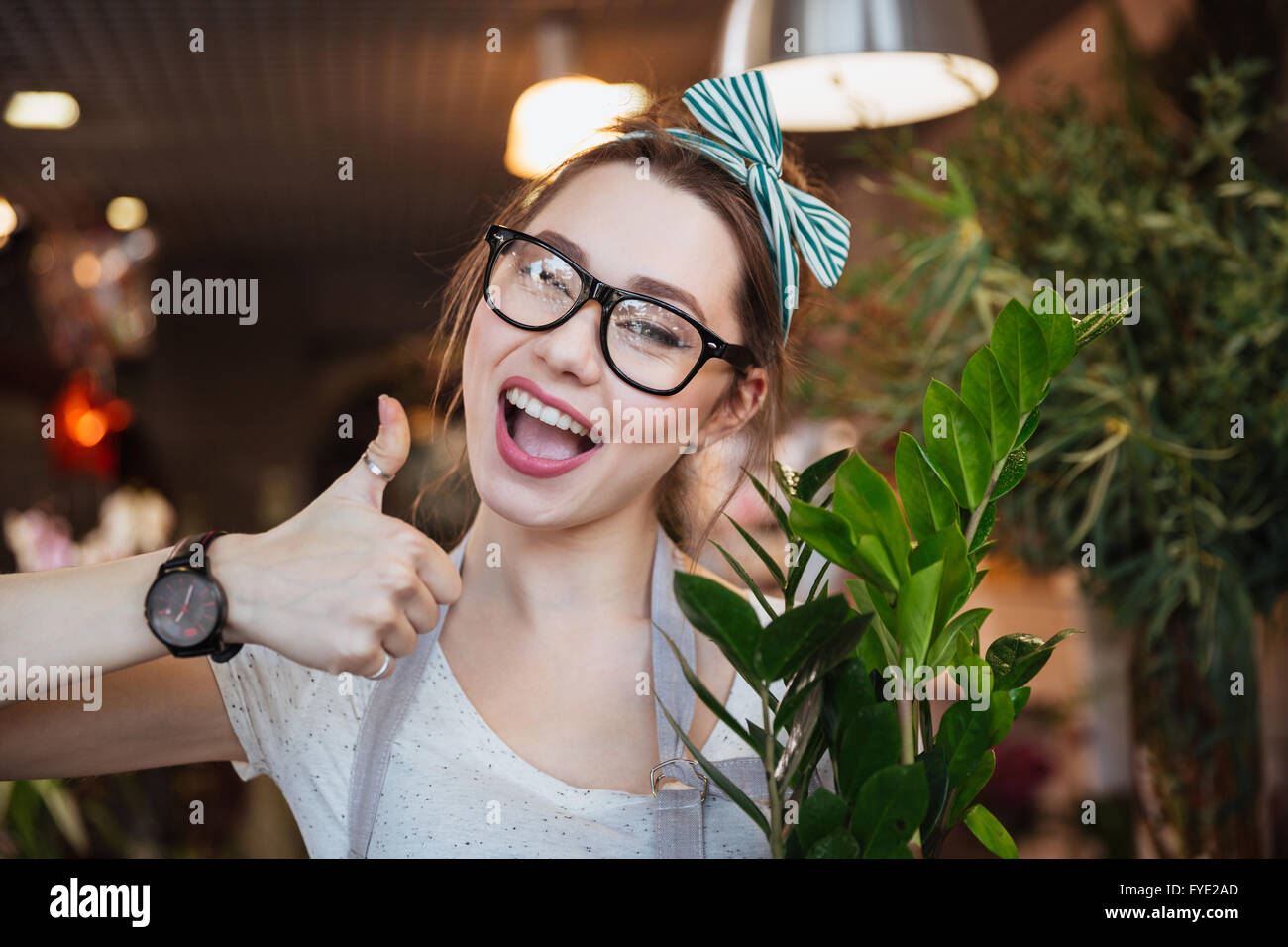 Happy charming young woman florist showing thumbs up in flower shop ...