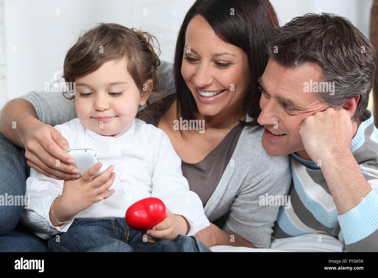 Toddler holding mobile telephone whilst sat with parents on sofa Stock Photo