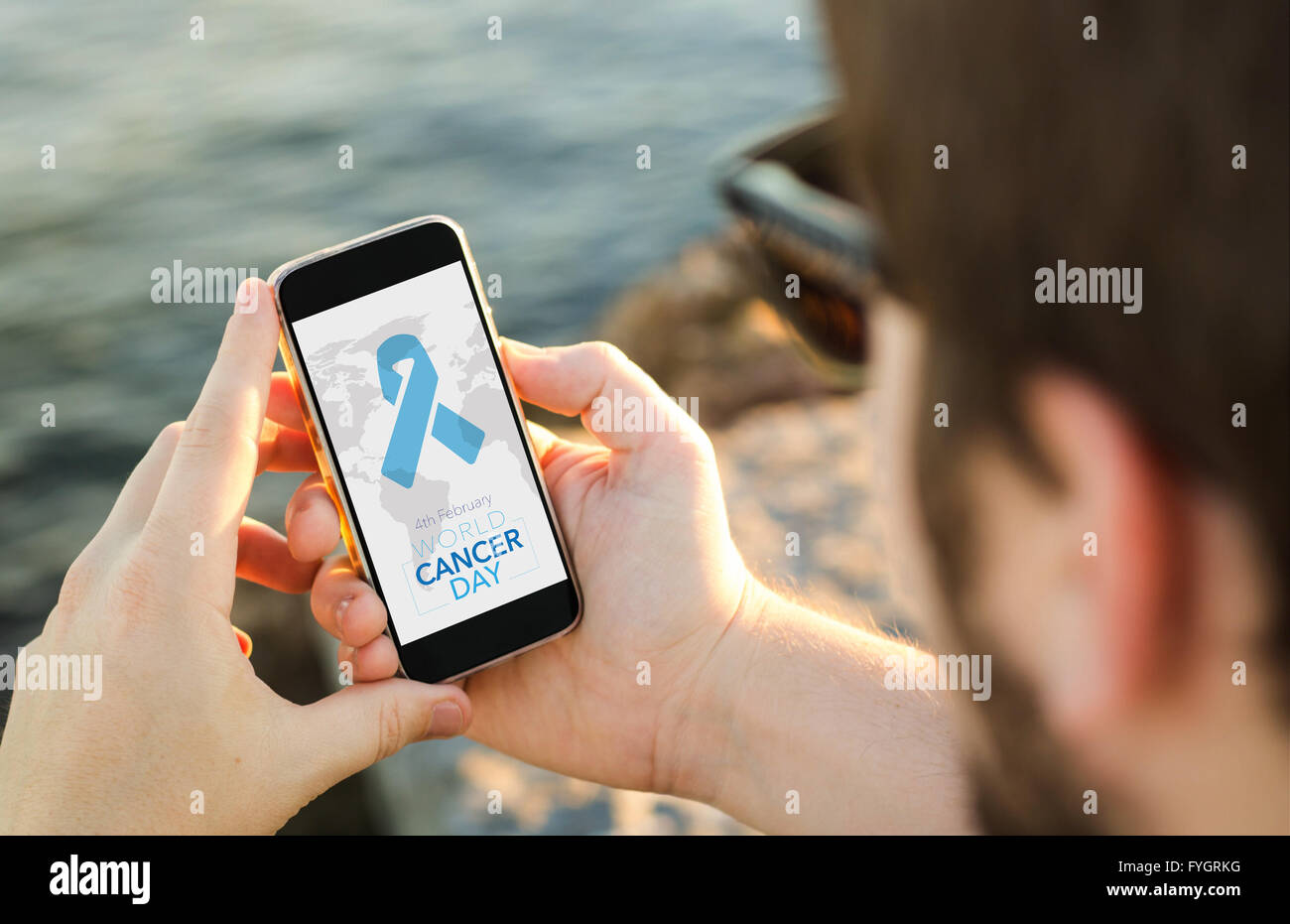 man on the coast using his smartphone to look a World Cancer Day graphic. All screen graphics are made up. Stock Photo