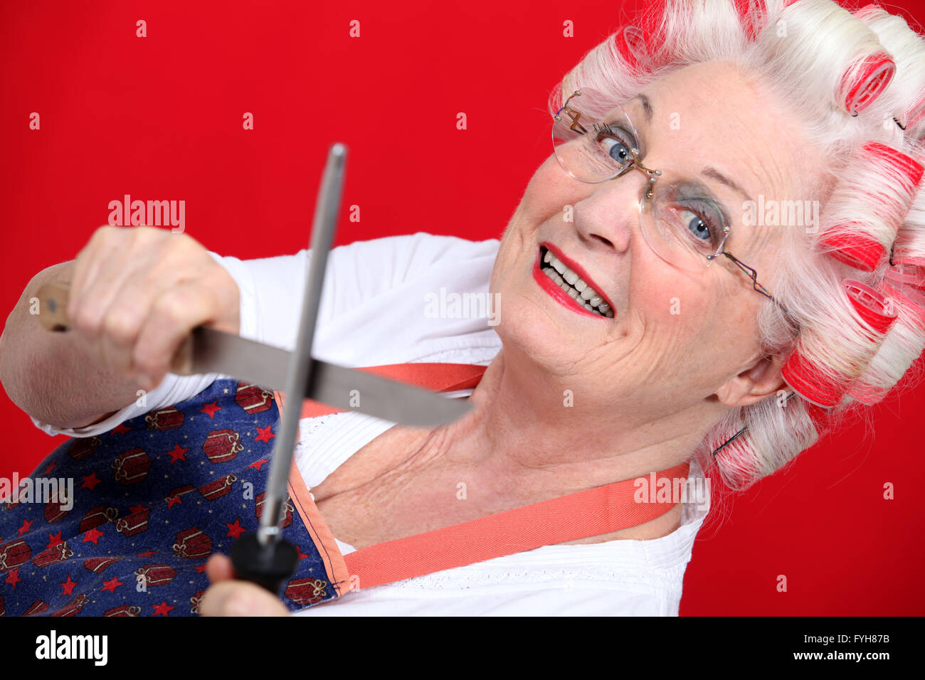 grandmother with haircurlers sharpening knives against red background Stock Photo
