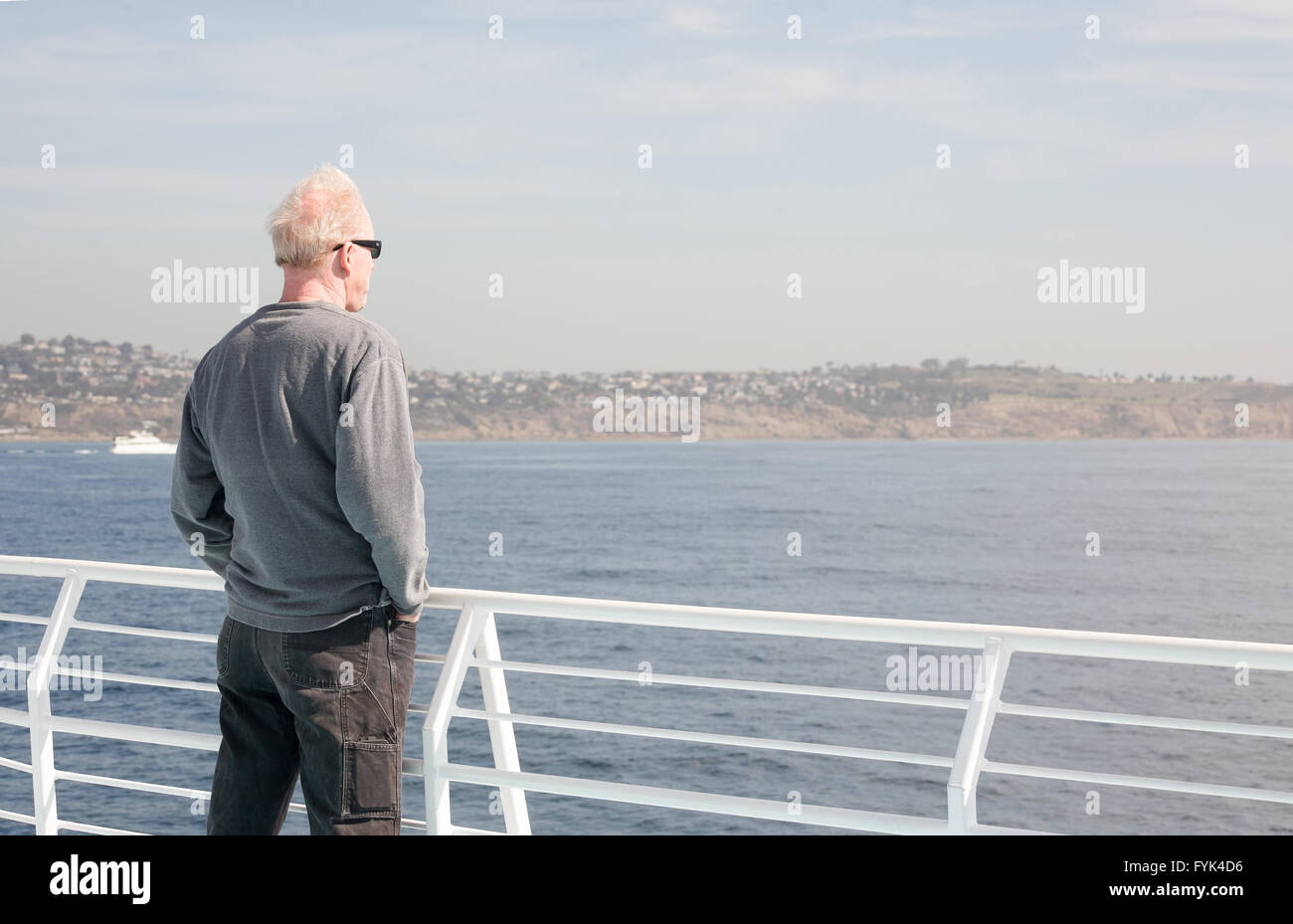 Middle age man dressed casually wearing sunglasses stands next to a white railing on a boat looking out to the water Stock Photo
