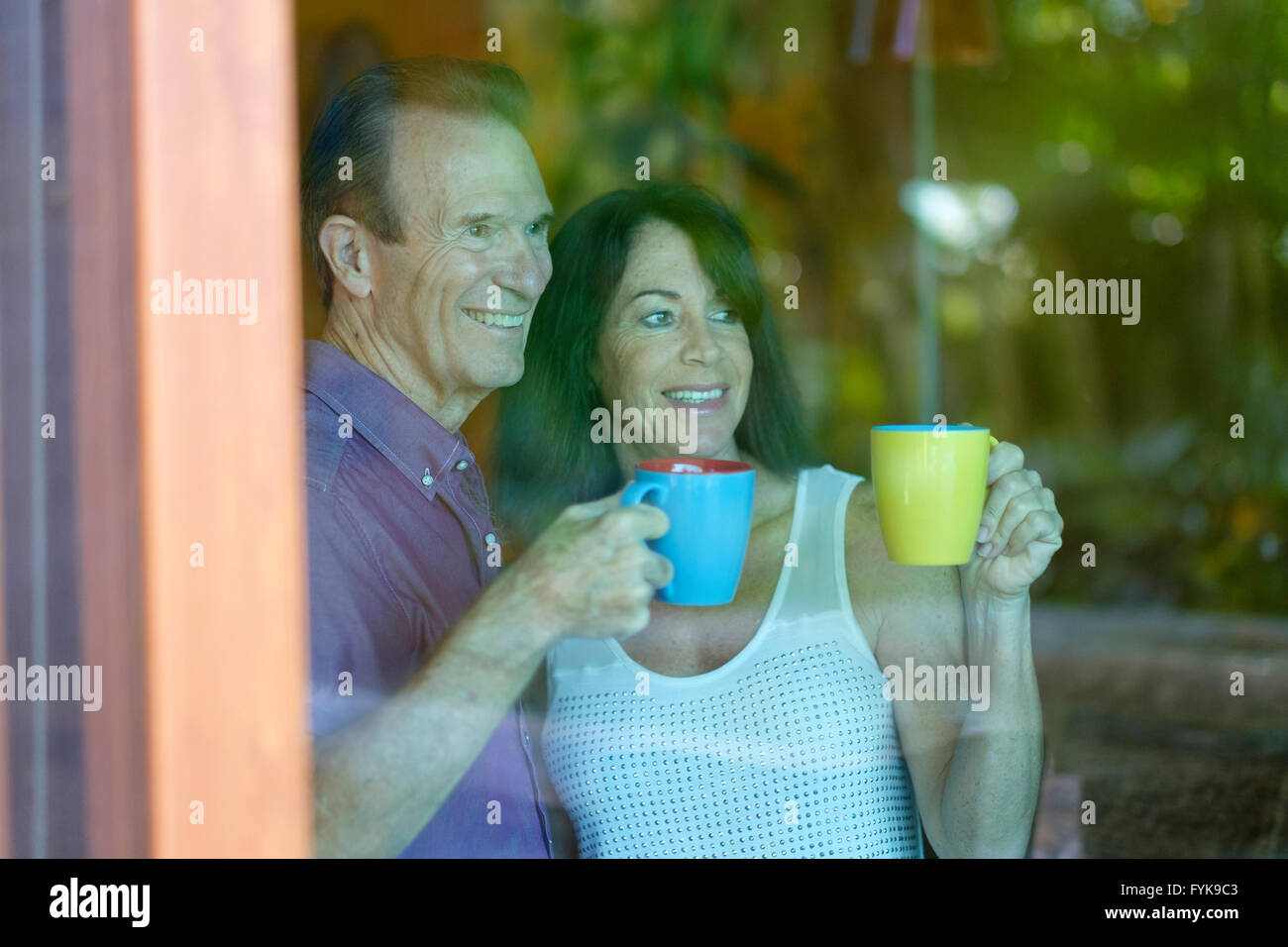 Senior man and woman looking out of the tinted glass doors of their home while drinking coffee from mugs Stock Photo