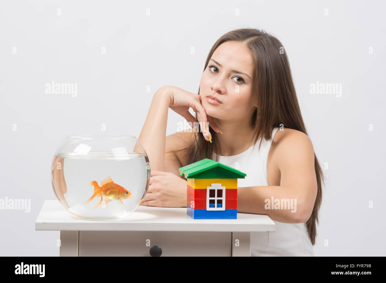 Thoughtful girl sitting at a table on which there is an aquarium with goldfish and toy house Stock Photo