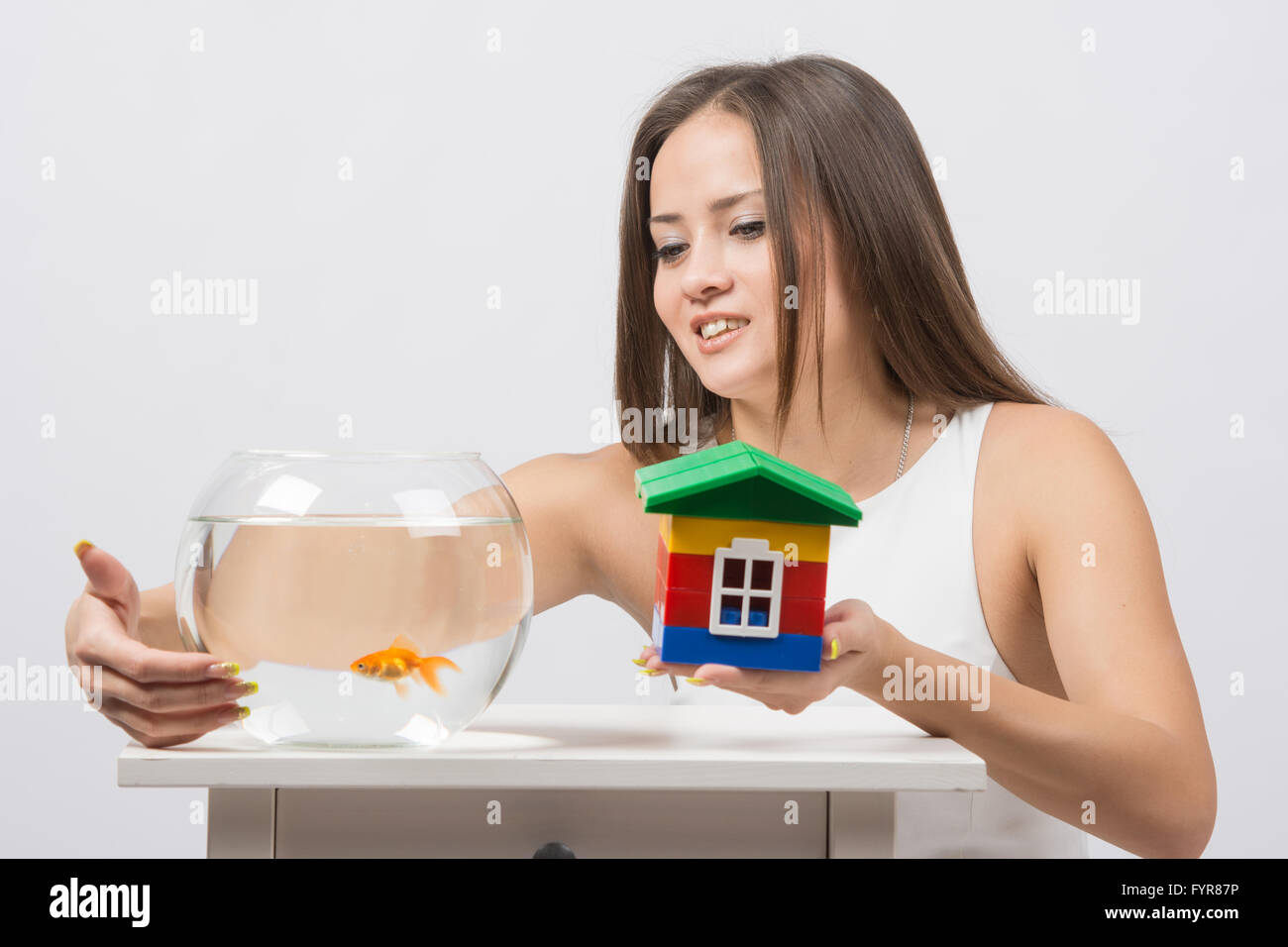 She knocks on the wall of the aquarium with goldfish and the other hand holding a toy house Stock Photo
