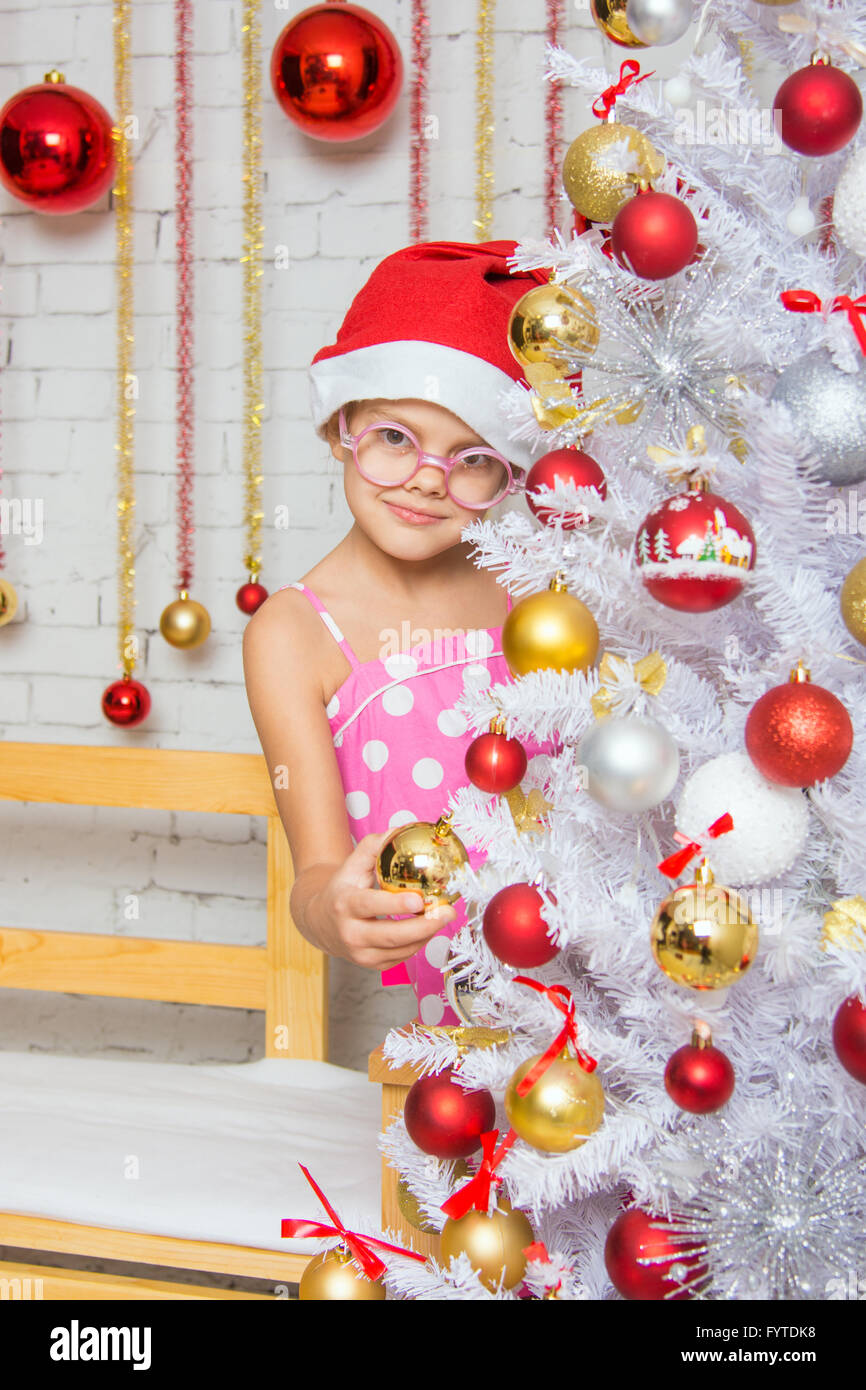 The girl in round glasses and a red Christmas hat hangs balls on a snowy New Years Christmas tree Stock Photo