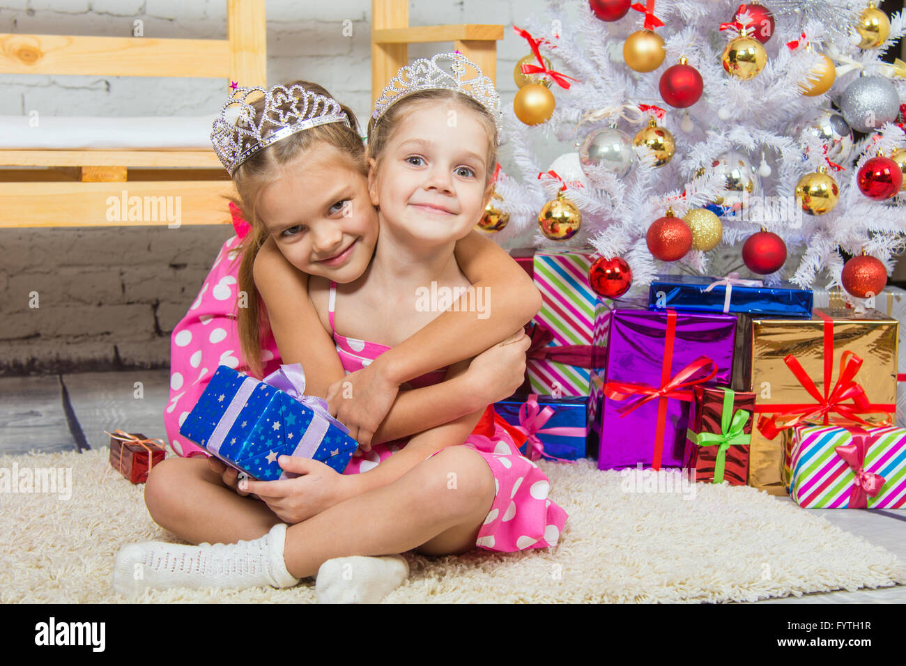 Girl hugging another girl sitting on a mat at the Christmas tree Stock Photo