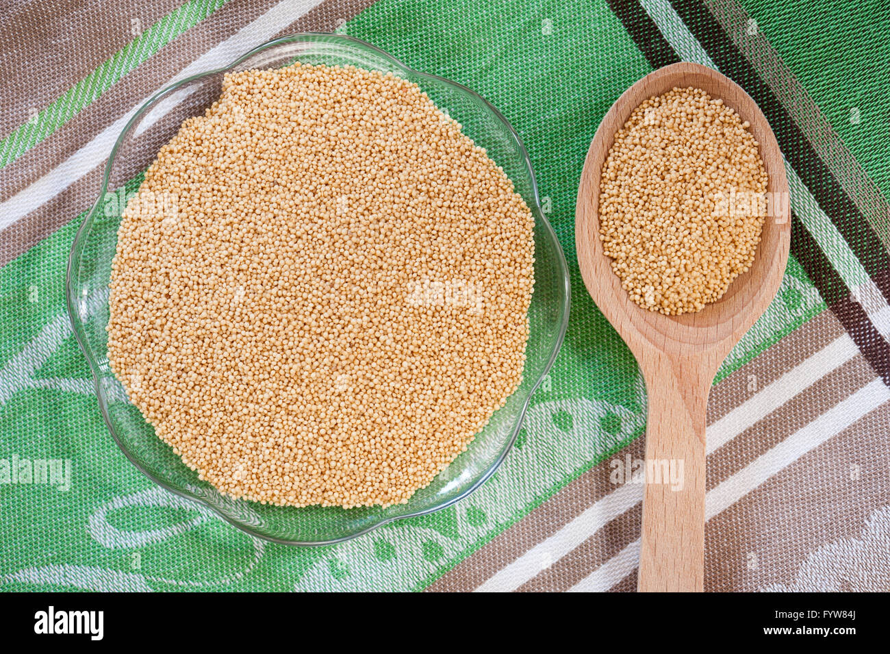 Tiny amaranth grains lying on glass saucer and wooden spoon on green cloth, raw product healthy food staple portion. Stock Photo