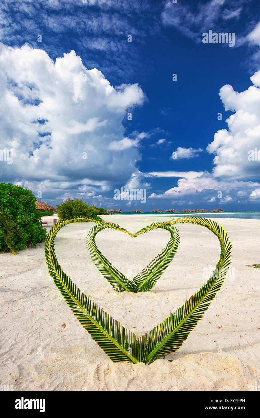 Heart made of palm tree leaves on tropical island with overwater bungalows in the background. Stock Photo