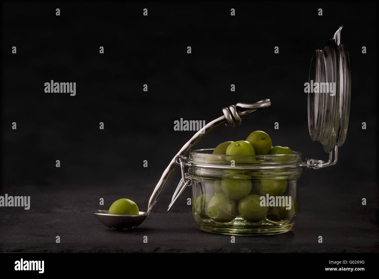 Jar of green olives with single olive on spoon against grey slate background. Stock Photo