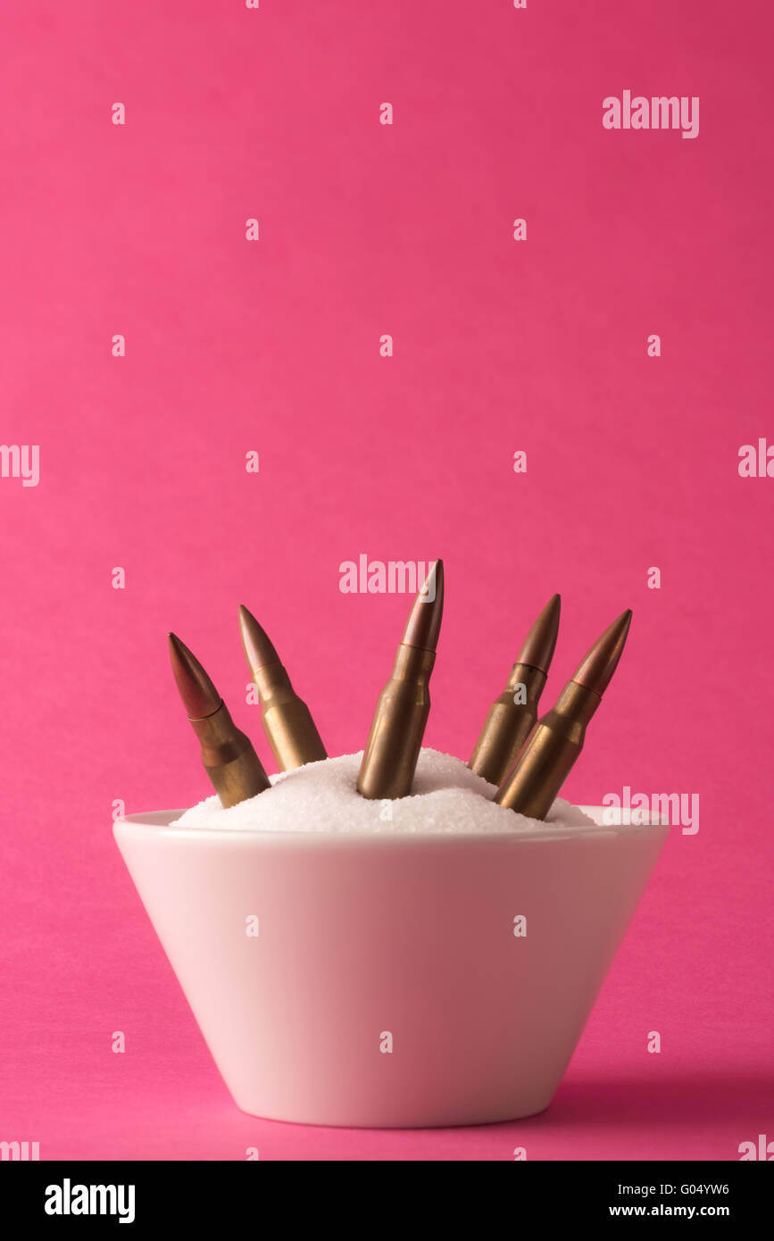 White bowl full of white sugar and rifle bullets against pink background Stock Photo