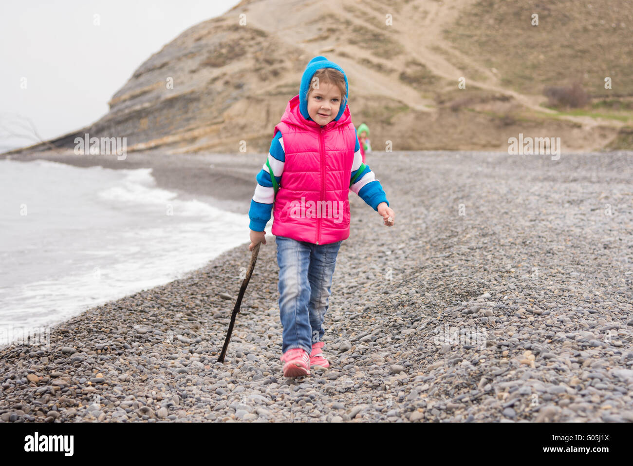 Five-year girl walking on the pebble beach in the warm bright clothes with a stick in his hand Stock Photo