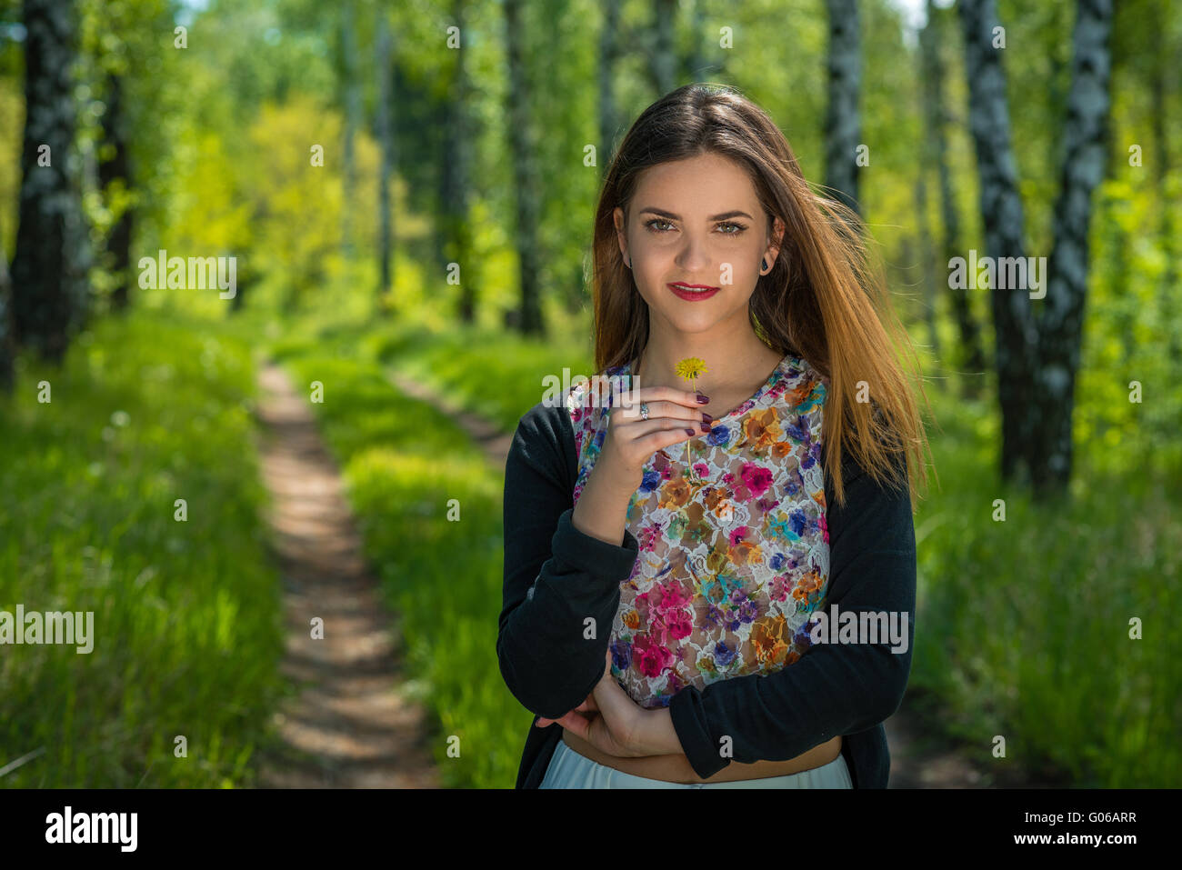 Young woman with red lips and brown hair is holding one yellow dandelion in hand while standing on a footpath in a forest during Stock Photo