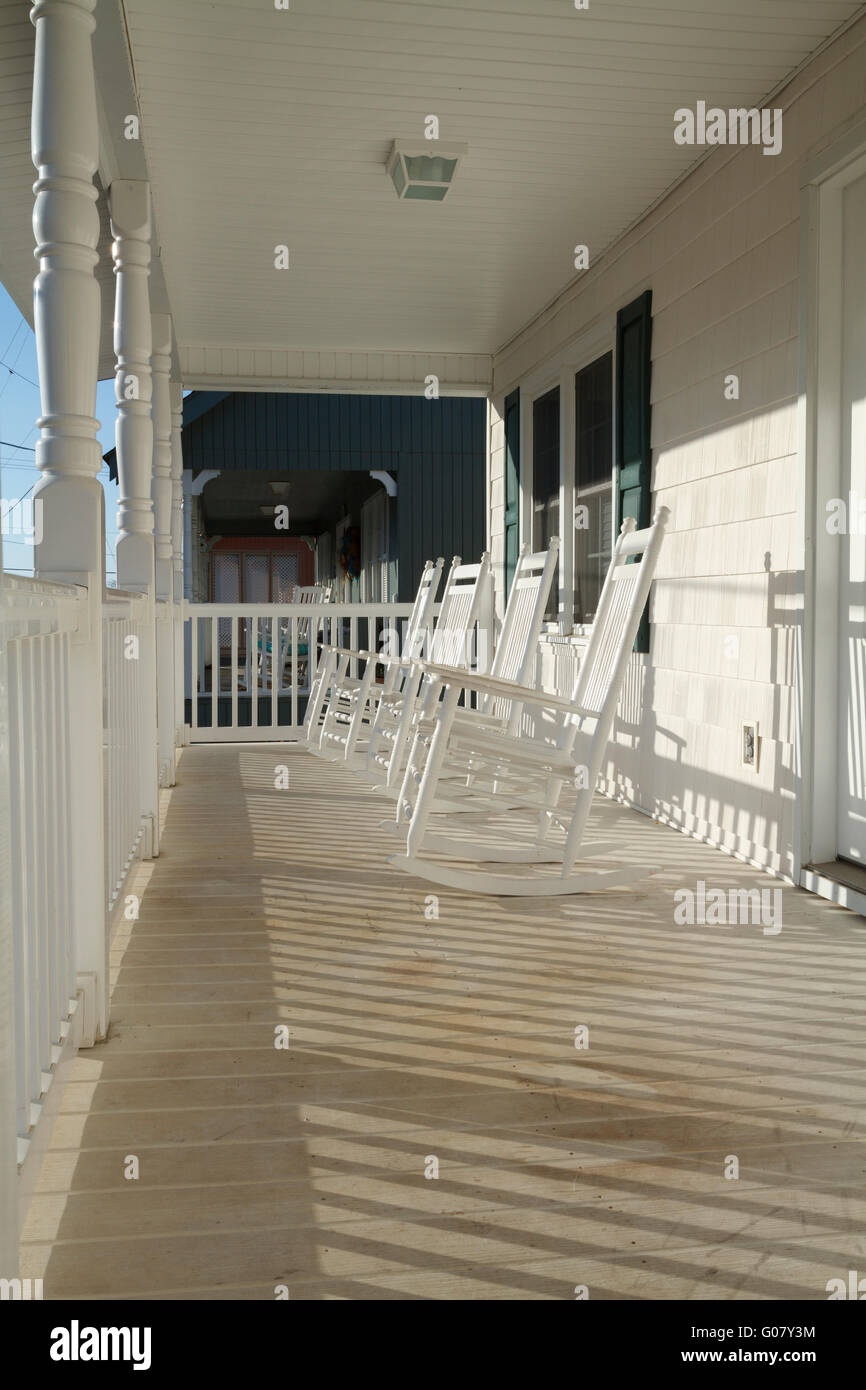 A row of four used and weathered white painted wooden country style rocking chairs on a worn porch at sunrise on a sunny day. Stock Photo