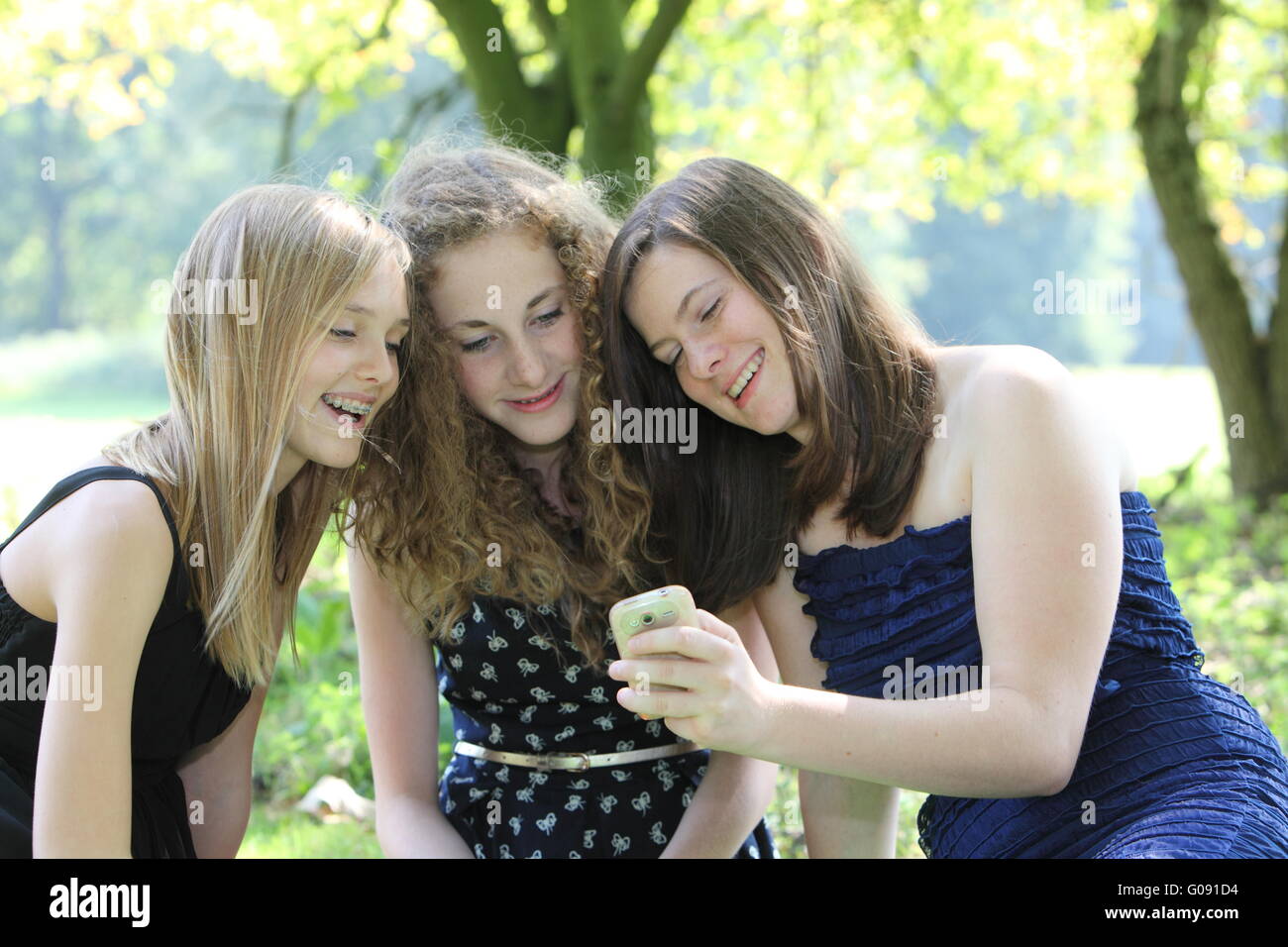 Three happy teenagers reading an sms on a mobile phone as they sit grouped together on a rug on the grass in a park while relaxi Stock Photo