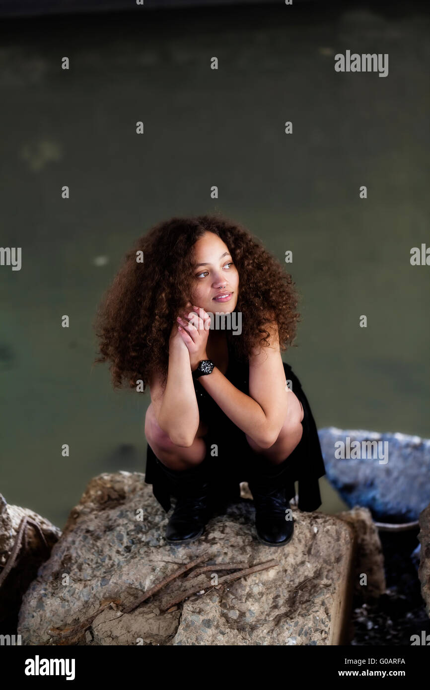 Mixed Heritage Woman Black Dress Squatting On Concrete Block Stock Photo