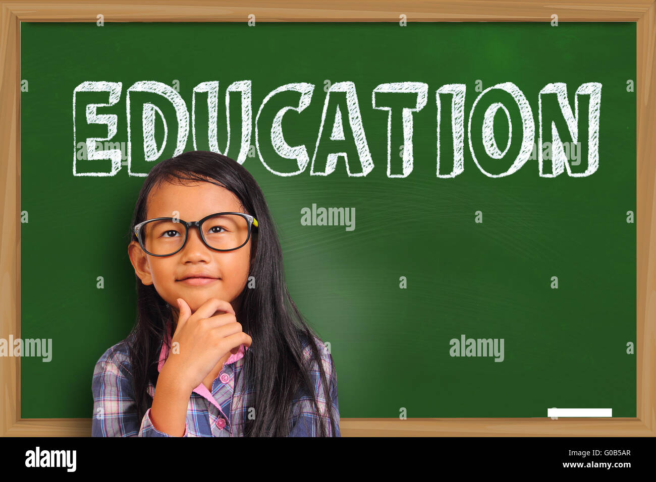 Little Asian student girl smiling and thinking over green chalkboard with education word written on it Stock Photo