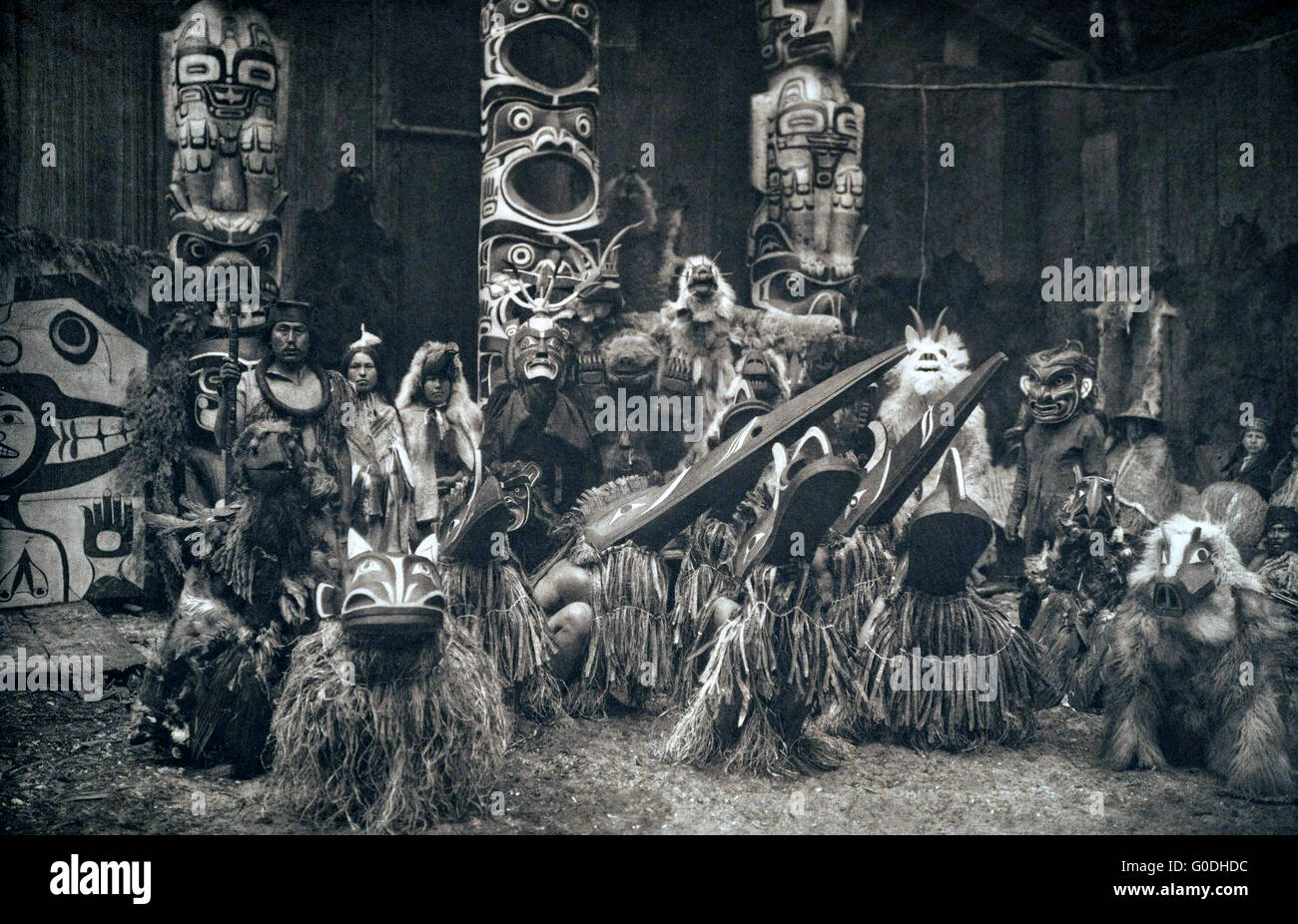 Native American Kwakiutl or Qagyuhi dancers wearing masks and costumes during a traditional potlatch 1914 in British Columbia, Canada. The chief on the far left holds a speaker's staff. Three totem poles in background. Stock Photo