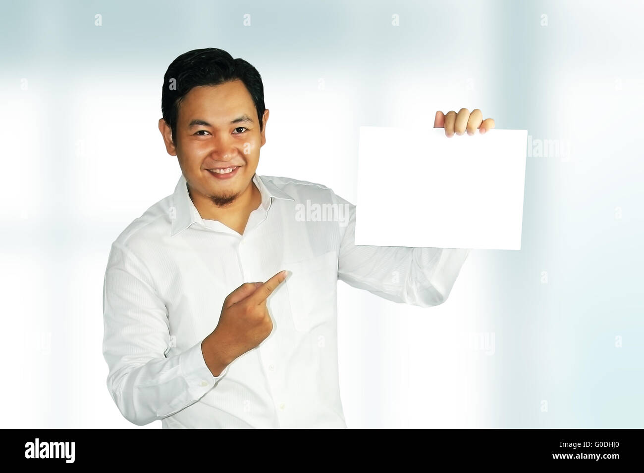 Image of a young Asian male smiling and holding white empty paper over bright background Stock Photo