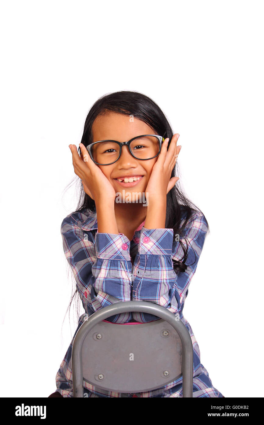 Cute little girl with glasses sitting backward on chair smiling and touching her cheek isolated on white Stock Photo