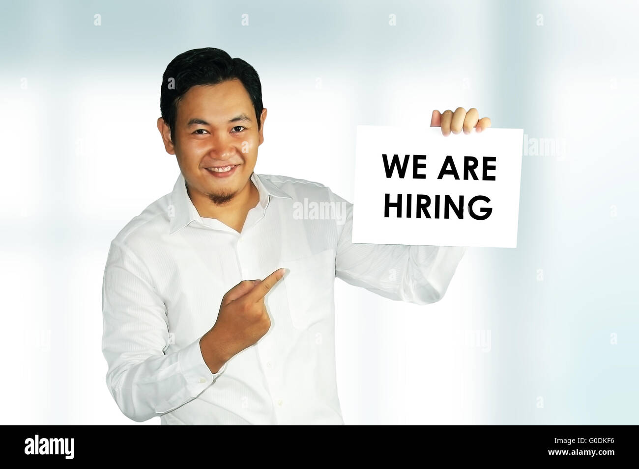 Image of a young Asian male holding white paper with We Are Hiring words written on it over bright background Stock Photo