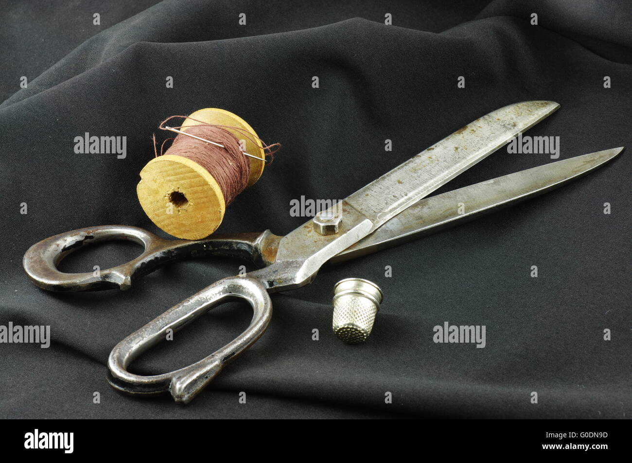 Old dressmaker shears, wooden spool of thread with a needle and a thimble. On a dark, undulating fabric. Stock Photo