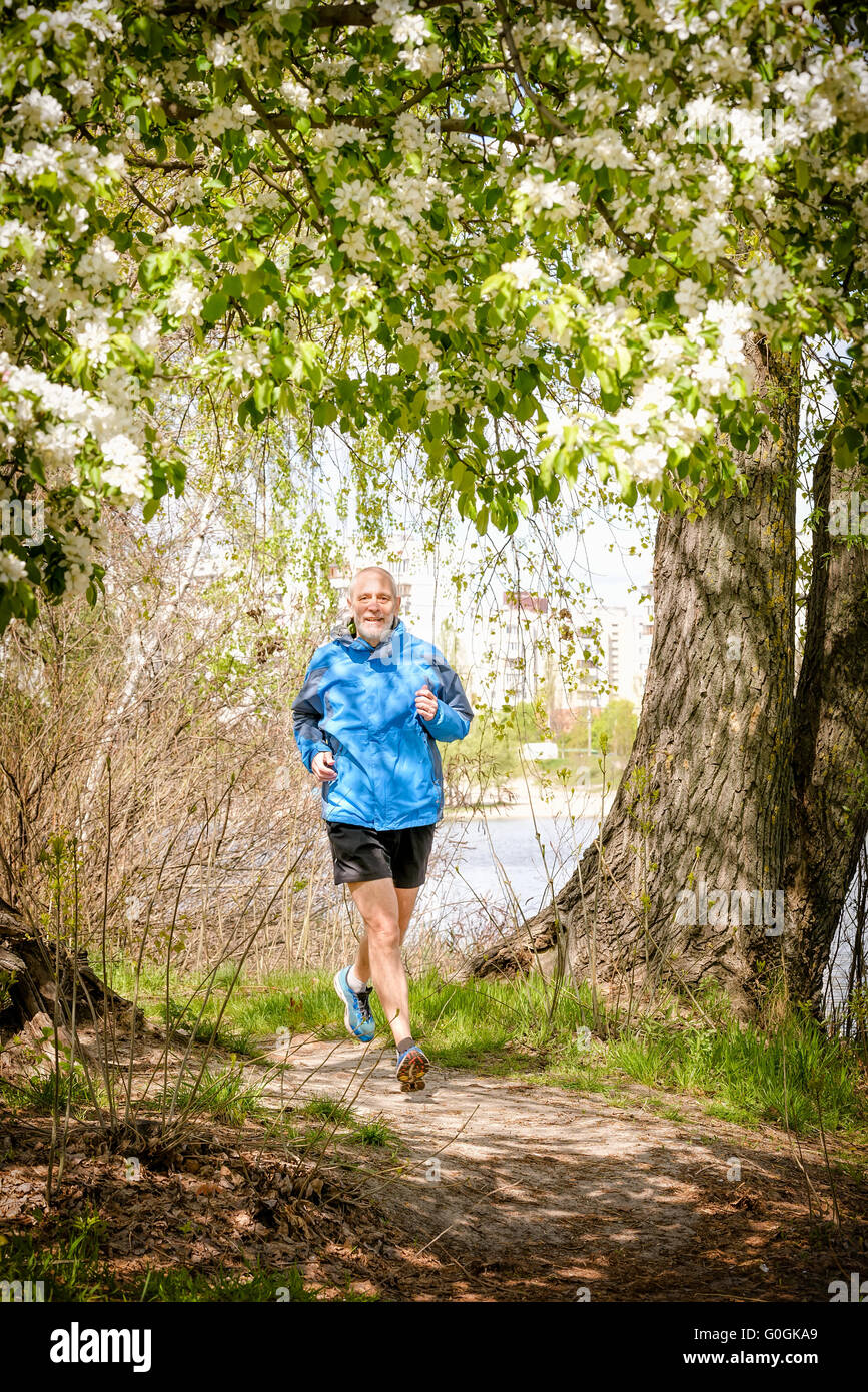 A senior man worn in black and blue is running in the forest, close to the lake, under an apple blossom, during a warm spring da Stock Photo
