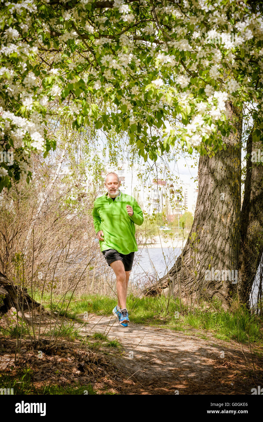 A senior man worn in black and green is running in the forest, close to the lake, under an apple blossom, during a warm spring d Stock Photo