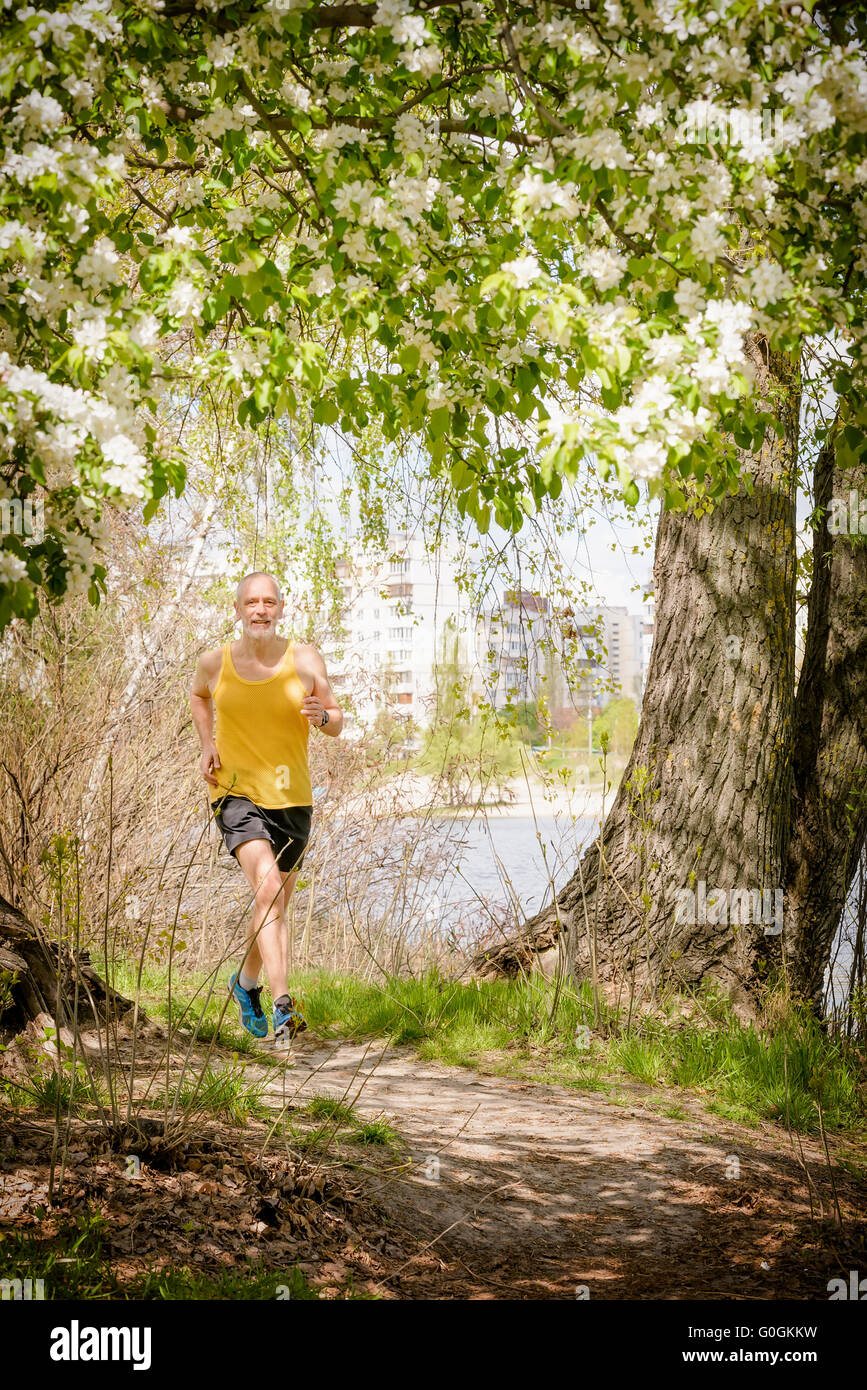 A senior man worn in black and yellow is running in the forest, close to the lake, during a warm spring day Stock Photo