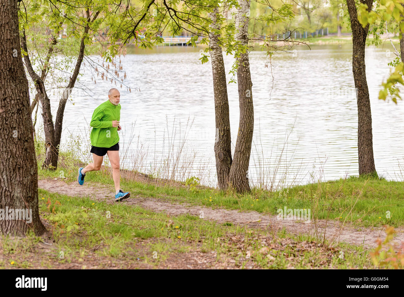 A tired senior man worn in black and green is running in the park, close to the lake, during a gray spring day Stock Photo