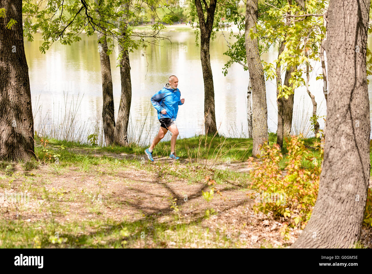 A senior man worn in black and blue is running close to the lake during a warm spring day Stock Photo