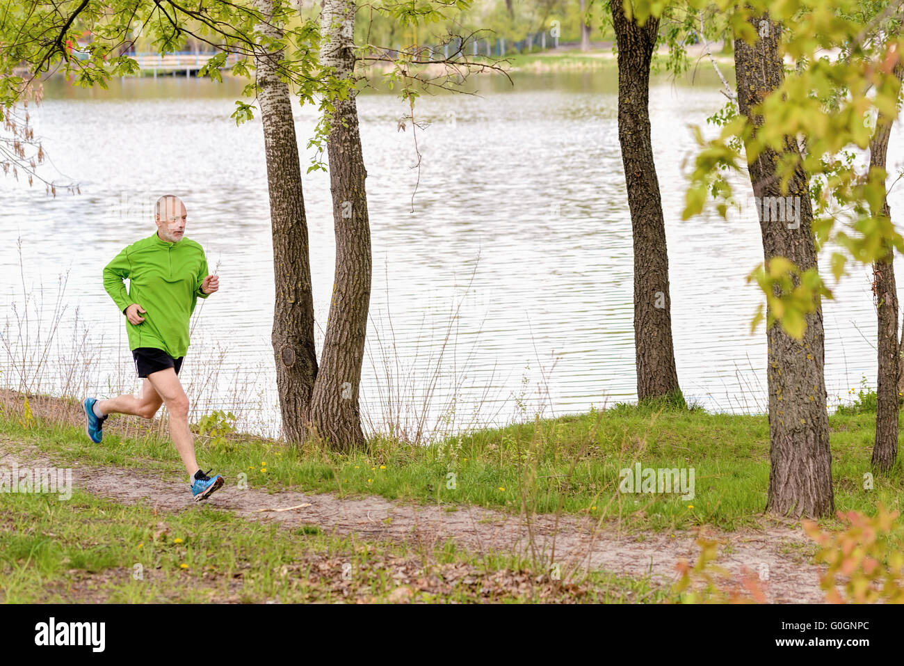 A senior man worn in black and green is running in the park, close to the lake, during a gray spring day Stock Photo